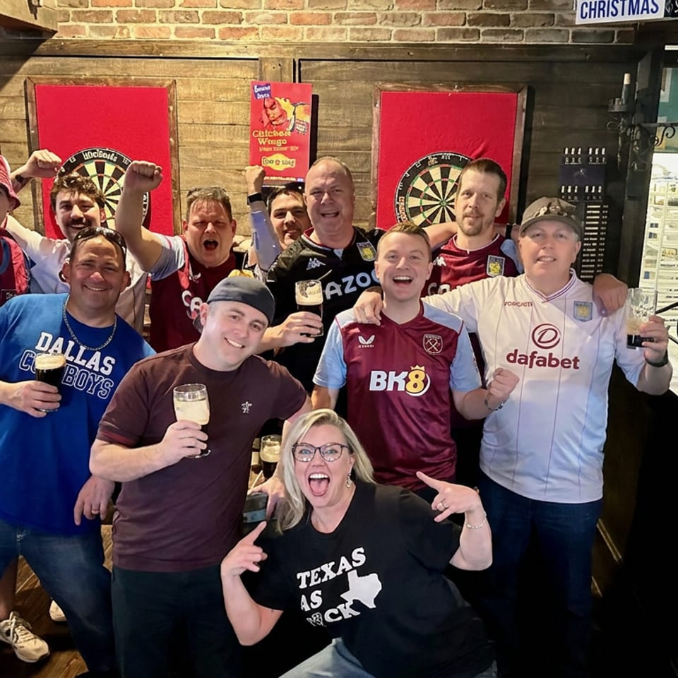 Group of smiling sports fans raising drinks and fists inside a lively pub with dartboards and team jerseys.