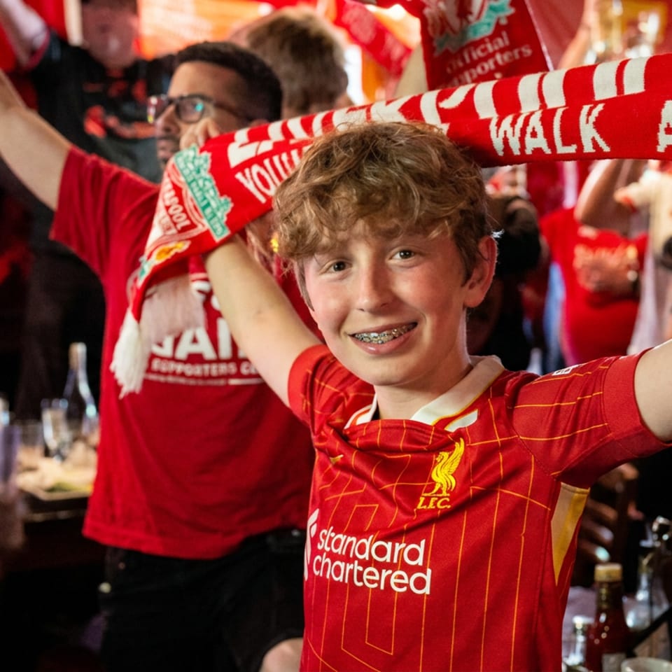 Young Liverpool football fan raises a scarf amid cheering supporters in a lively restaurant or pub.