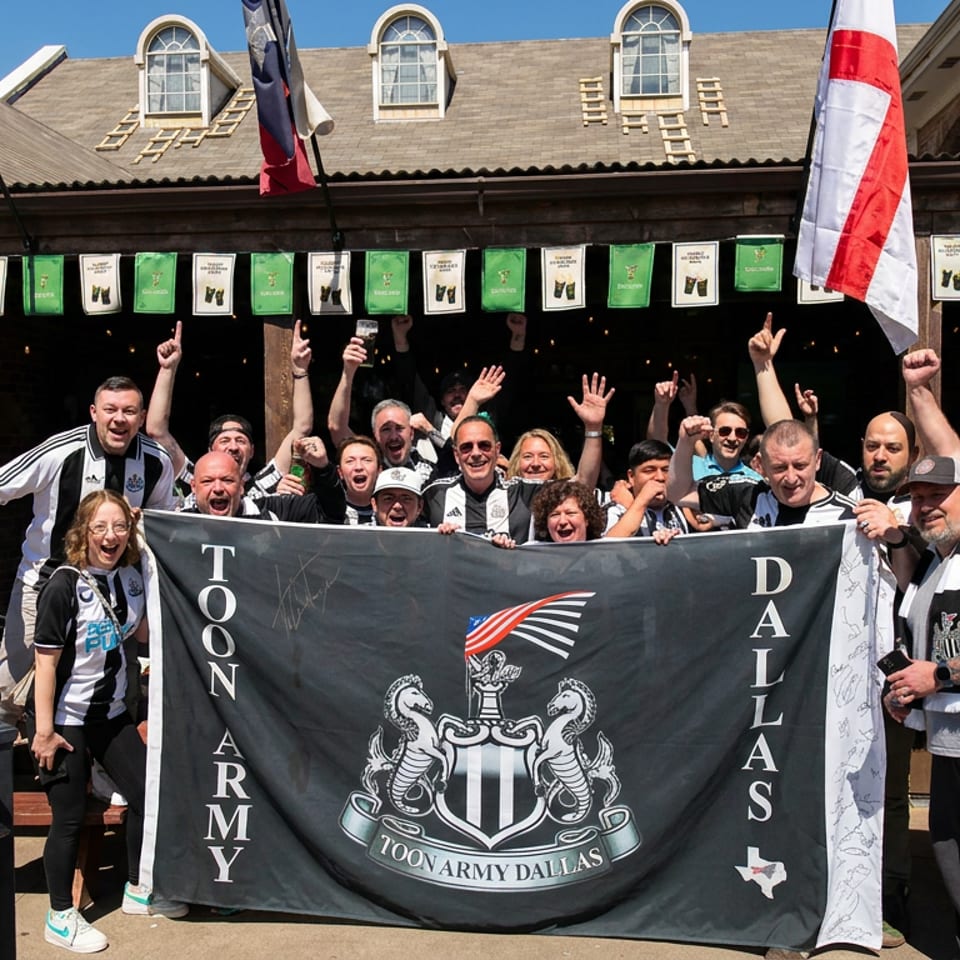 Toon Army Dallas supporters cheer outside a brick pub, holding a large signed banner beneath American and club flags.