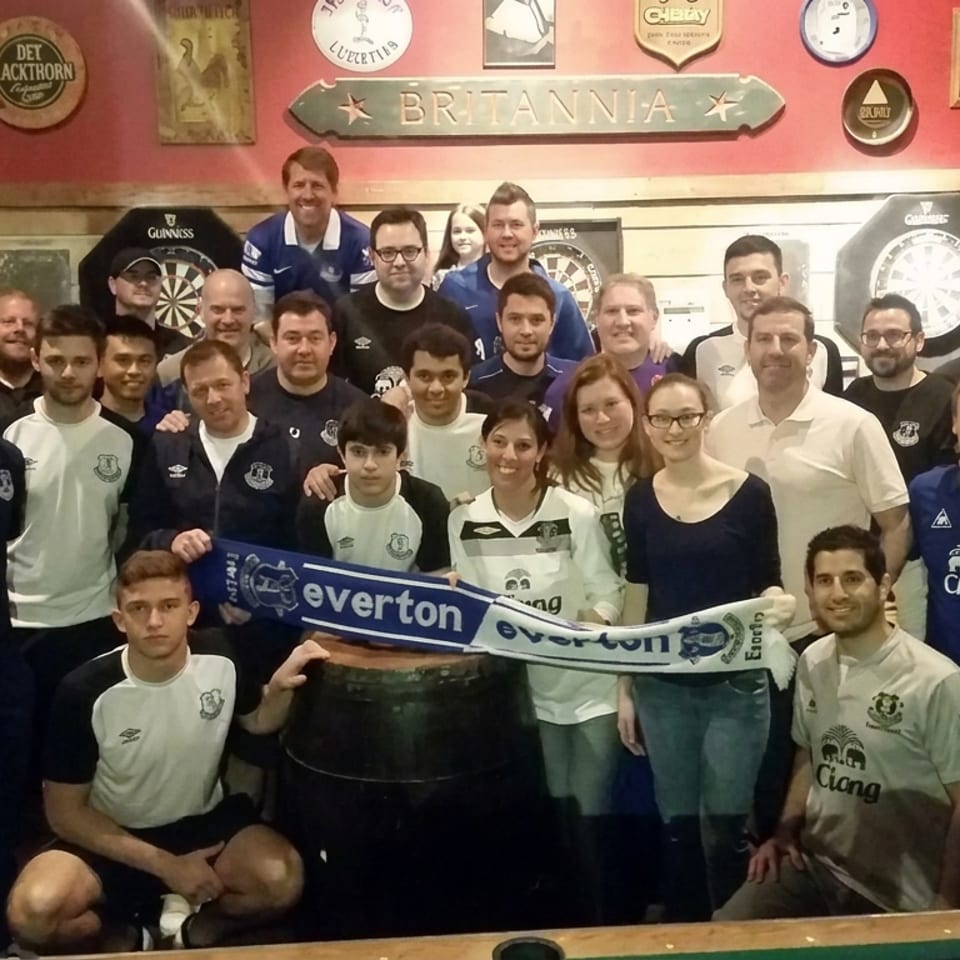 Group of Everton supporters posing with a scarf inside a Britannia-themed pub decorated with dartboards and memorabilia.