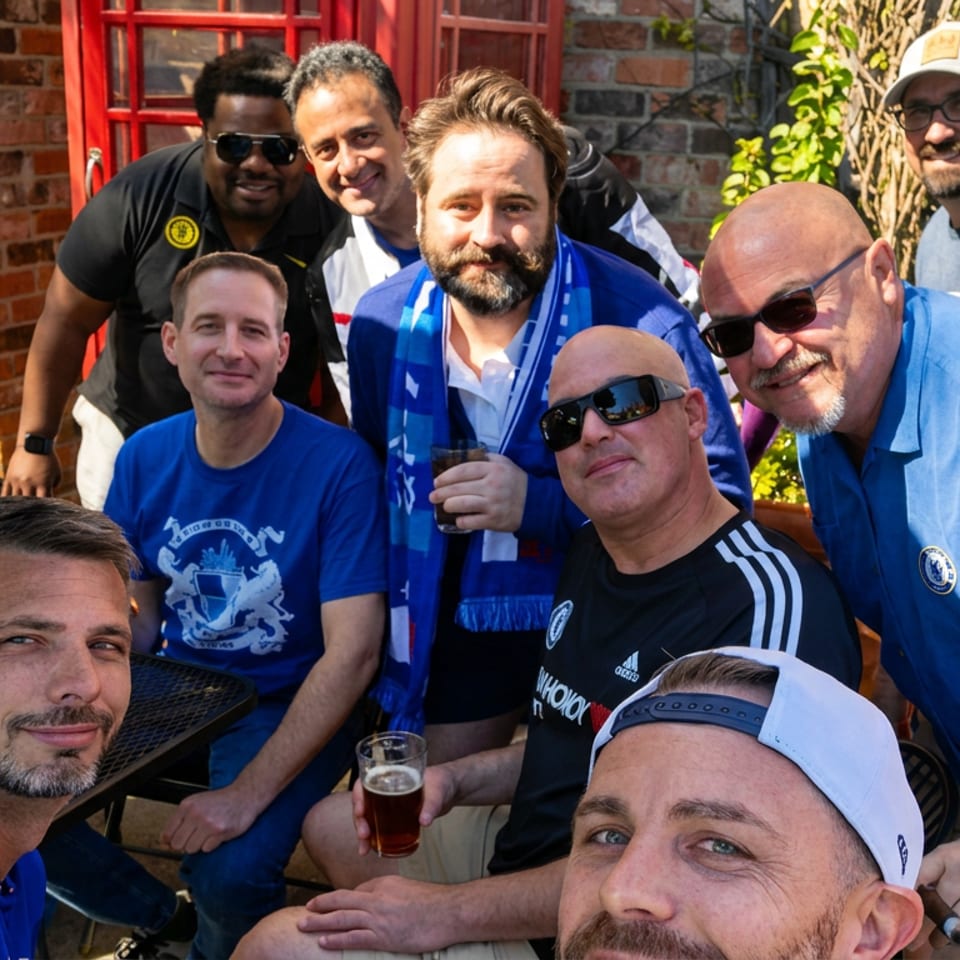 Group of soccer fans posing with drinks on a sunny restaurant patio outside a brick building.
