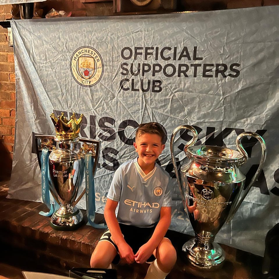 Young Manchester City supporter beside trophies in front of the Frisco Sky Blues supporters club banner.