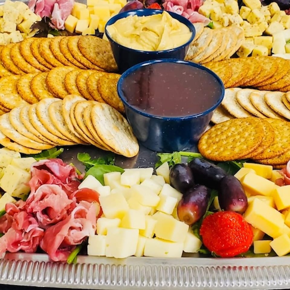 Cheese platter with crackers, sliced meats, grapes, strawberries, and dips.