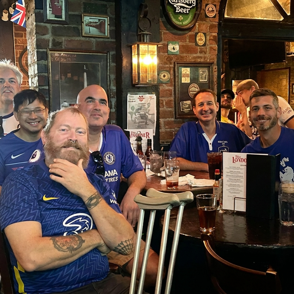 Chelsea football supporters pose together around a pub table surrounded by drinks, brick walls, and sports memorabilia.