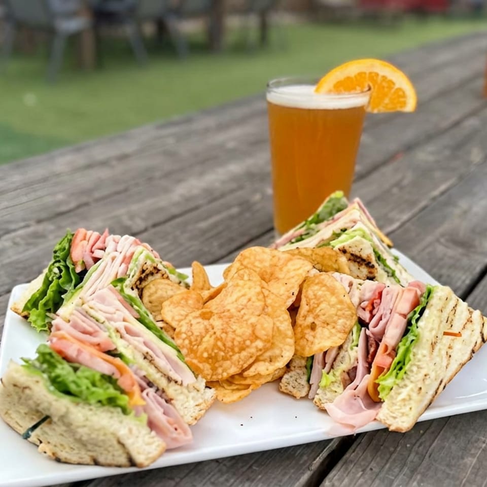 Plate of ham and turkey sandwiches with lettuce, tortilla chips, and an orange-garnished beer on a wooden picnic table.