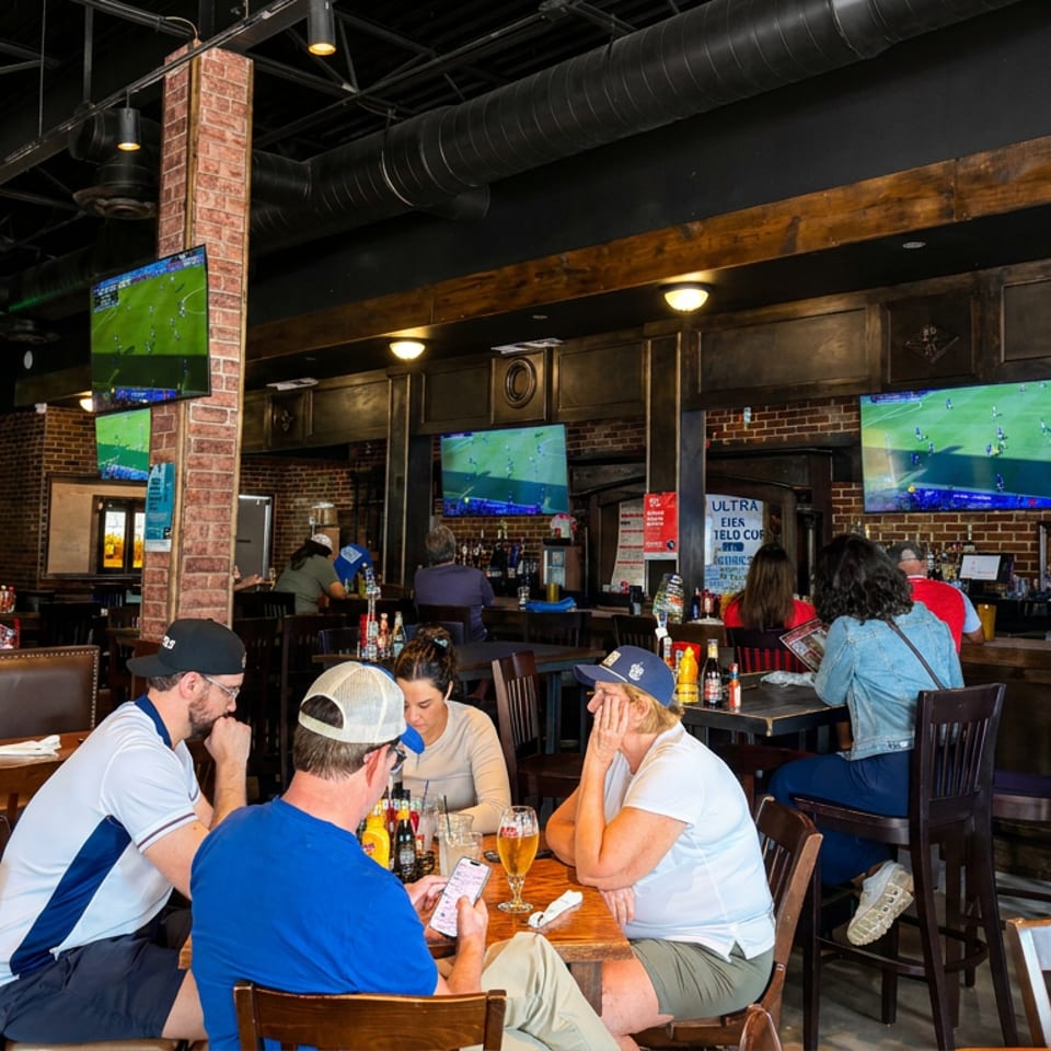Busy sports bar with exposed brick, multiple televisions showing soccer, and patrons dining at tables with drinks.
