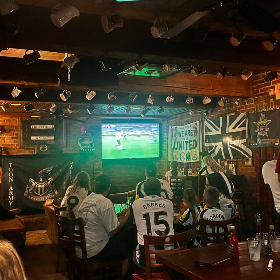 Crowded sports pub with fans watching soccer on television, surrounded by football banners, memorabilia, tables, and drinks.