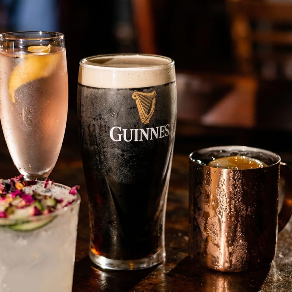 Three drinks on a dimly lit bar: Guinness stout, lemon-garnished sparkling water, and a copper mug beside a floral cocktail.