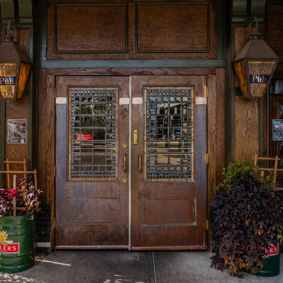 Rustic pub entrance with wooden double doors, lanterns, potted plants, and green Miller beer barrels outside.