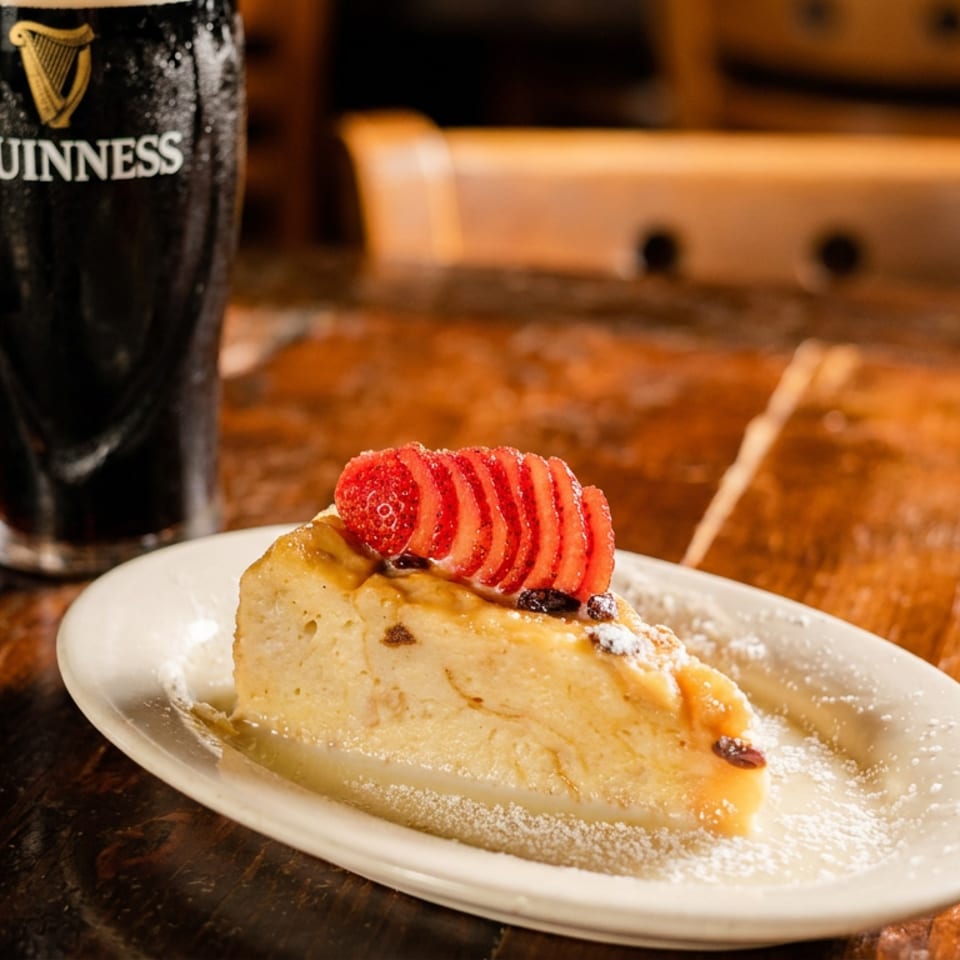 Slice of fruit-topped cheesecake dusted with powdered sugar on a plate beside a pint of Guinness at a wooden pub table.