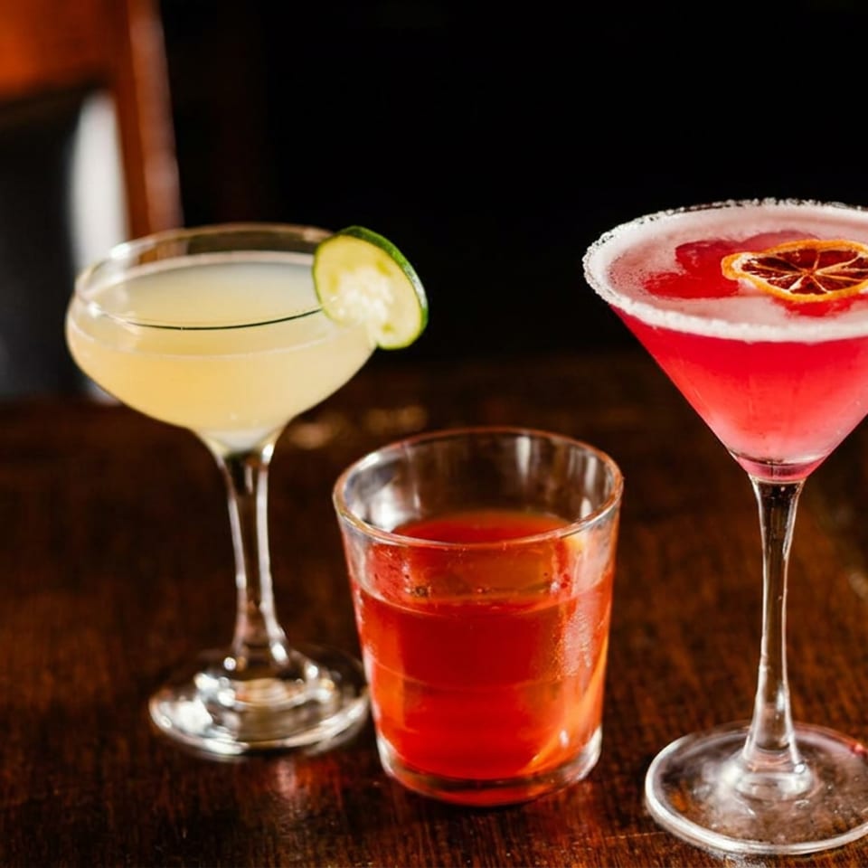 Three colorful cocktails sit on a dark wooden table, including a lime-garnished pale drink, red tumbler, and citrus-garnished martini.